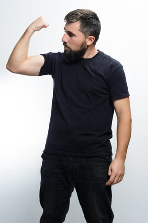 A man with a beard, wearing a dark shirt and jeans, flexes his bicep against a plain white background, looking at his arm.の写真素材