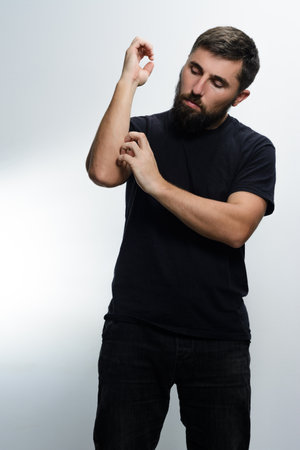 A man with a beard, wearing a black shirt and dark pants, scratches his arm against a plain white backdrop. He appears to be experiencing discomfort or irritation.の写真素材
