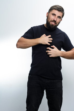 A man with a beard, wearing a black shirt and jeans, scratches his chest with both hands. He appears uncomfortable, suggesting skin irritation or allergy.の写真素材