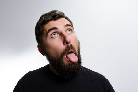A bearded man looks upwards with his tongue sticking out against a plain background. He appears to be making a silly or playful expression.の写真素材