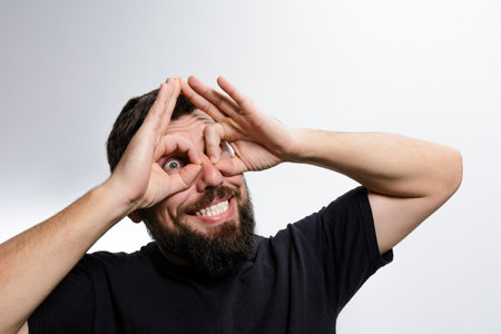 A bearded man with a playful expression forms binoculars around his eyes using his hands, smiling broadly against a light background. His playful gesture conveys a sense of fun and lightheartedness.の写真素材