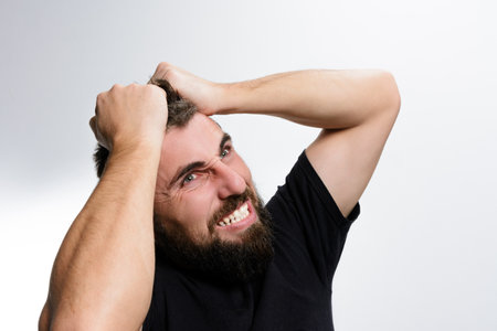 A bearded man grips his hair tightly, his face contorted in a grimace of frustration and anger. The image portrays stress, anxiety, and emotional distress against a plain background.の写真素材