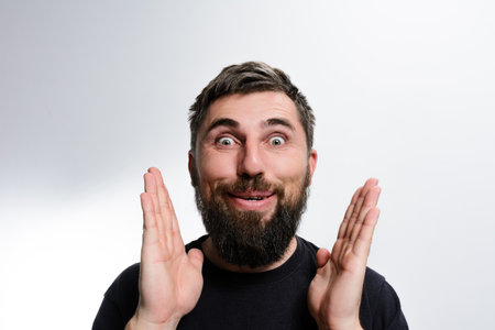 A bearded man expresses surprise with wide eyes and raised hands against a light background. His joyful expression conveys astonishment and excitement.の写真素材