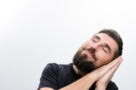 A bearded man with closed eyes rests his head on his hands, simulating sleep against a white background, expressing tranquility and rest.の写真素材