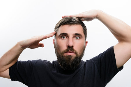 A bearded man in a dark shirt salutes with one hand while adjusting his hair with the other against a white background. His expression is serious.の写真素材