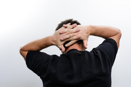 Rear view of a man with his hands clasped behind his head against a plain background. He's wearing a dark t-shirt.の写真素材