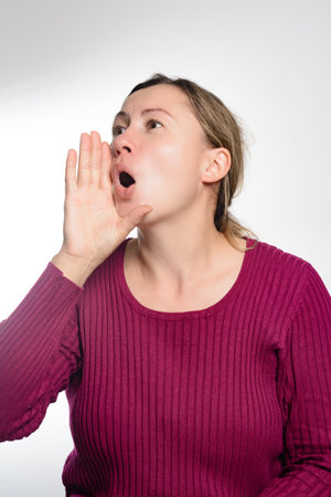 A middle-aged woman with light brown hair, wearing a dark pink ribbed sweater, cups her hand to her...