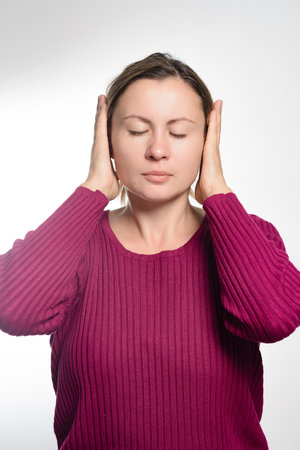 A woman wearing a dark pink sweater stands against a white backdrop, her eyes closed and hands covering her ears, conveying a sense of peace, quiet, or blocking out noise.の写真素材