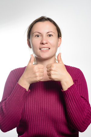 A woman in a maroon sweater smiles brightly at the camera while giving two thumbs up against a white background, expressing positivity and approval.の写真素材