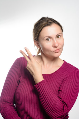 A woman in a maroon sweater holds up two fingers against a white background, her expression slightly playful and inquisitive.の写真素材