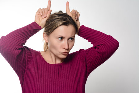 A woman with a playful expression makes horns with her fingers above her head. She wears a ribbed, dark pink sweater against a light background.の写真素材