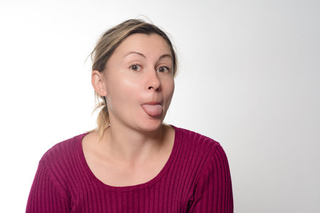 Headshot of a woman with a playful expression, sticking her tongue out against a plain background. She wears a maroon ribbed sweater and has her hair tied back.の写真素材