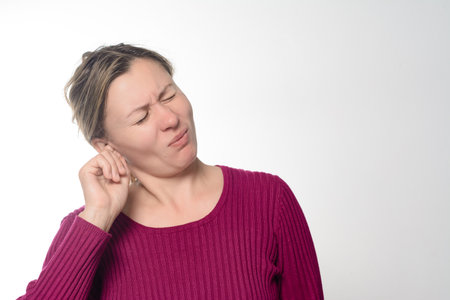 A woman with her eyes closed, touching her ear, conveying discomfort or pain. She is wearing a maroon sweater against a plain background, suggesting potential earache or other auditory issue.の写真素材