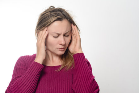 A woman with her eyes closed, wearing a maroon sweater, massages her temples with her fingers, expressing discomfort or pain, possibly due to a headache or stress.の写真素材