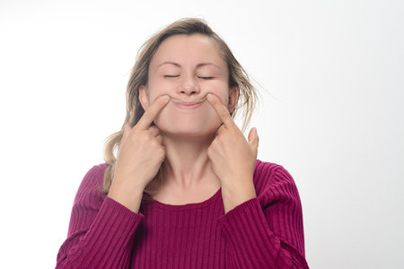 A woman with closed eyes uses her index fingers to push the corners of her mouth upwards in a forced or fake smile against a white background.の写真素材