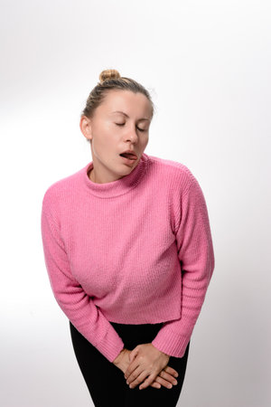 A woman with her hair in a bun, wearing a pink sweater and black pants, stands against a white background, her eyes closed, tongue slightly out, and hands clasped over her stomach.の写真素材