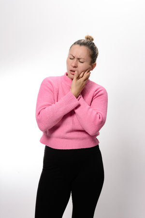 A woman in a pink sweater and black pants holds her jaw, her face expressing discomfort and pain, suggesting a toothache or dental issue against a white background.の写真素材
