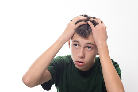 A young man with dark hair looks concerned, his hands on his head. He's wearing a dark green shirt. The background is plain white, focusing attention on his expression.の写真素材