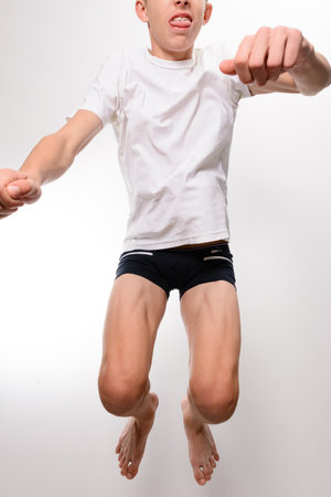 A low-angle shot of a teenage boy mid-jump against a light gray background. He's wearing a white t-shirt and dark shorts. The image captures his dynamic movement and youthful energy.の写真素材