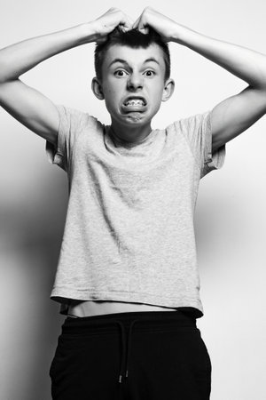 A black and white portrait of a teenage boy with a frustrated expression, hands in his hair. He's wearing a simple gray t-shirt and dark pants.の写真素材