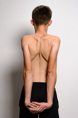 Rear view of a shirtless adolescent boy with his arms clasped behind his back. He's wearing dark athletic shorts. The image is simple and clean, ideal for health, fitness, or medical contexts.の写真素材