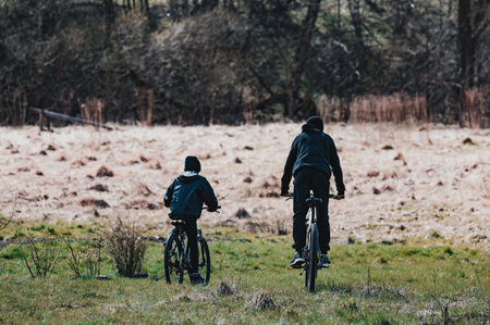 Two figures on bicycles ride across a grassy field, enjoying a peaceful outdoor adventure. The background features a blurred woodland and dry grass.の写真素材