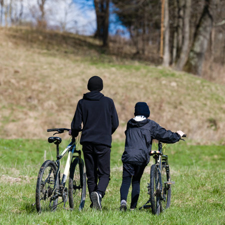 Two young people, seen from behind, walk along a grassy hill with their bicycles. They're dressed warmly and the setting suggests a spring or autumn day.の写真素材