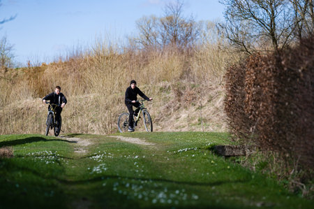 Two young people cycle along a country path, enjoying the springtime sunshine. The path winds through a field, past bushes and trees, creating a peaceful and scenic outdoor experience.の写真素材