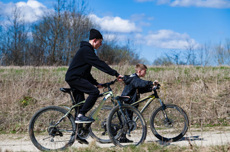 A man and boy cycle together on a sunny day. The path is dirt, and dry grass borders the road. They are enjoying the fresh air and sunshine.の写真素材