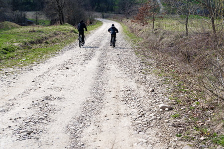 Two figures on bicycles travel along a rural, gravel road. The path winds through a landscape of sparse trees and grassy fields. The scene suggests a peaceful countryside setting.の写真素材