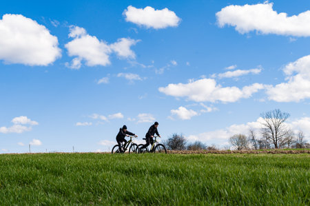 A picturesque scene of two individuals cycling across a vibrant green field. The clear blue sky is adorned with fluffy white clouds, creating a serene and peaceful atmosphere.の写真素材