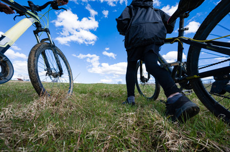 Low-angle view of two individuals pushing their mountain bikes across a grassy hill. The sky is bright blue with fluffy white clouds. A tranquil outdoor scene.の写真素材