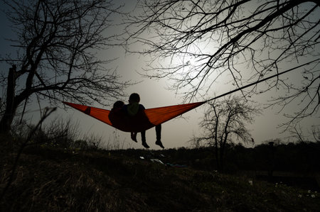 Silhouette of two people lounging in an orange hammock strung between leafless trees. Peaceful...