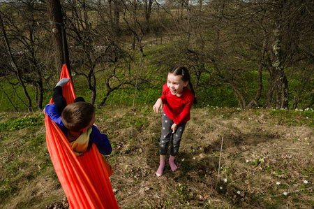 Two children enjoying playtime outdoors. One lies in a vibrant orange hammock, while the other stands, laughing. The setting is a sunny, grassy area with trees.の写真素材