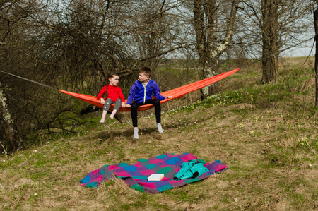 Siblings unwind in a vibrant orange hammock suspended between trees. A picnic blanket and jacket lie nearby, suggesting a relaxing outdoor excursion. The peaceful woodland setting enhances the scene.の写真素材