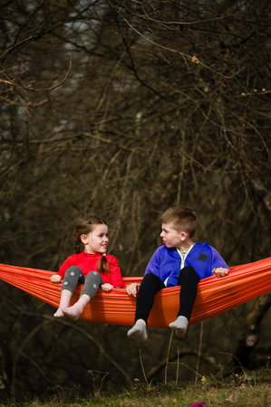 A girl and boy enjoy a leisurely afternoon together, sharing conversation in a bright orange hammock suspended between trees. The natural setting offers a tranquil backdrop.の写真素材