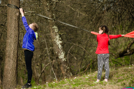 Two children, one in blue and one in red, playfully set up a rope between trees in a wooded area....