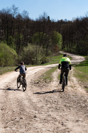 Two figures on bicycles ride along a winding dirt path. Trees and greenery line the road, creating a picturesque setting. A sunny day enhances the outdoor activity.の写真素材