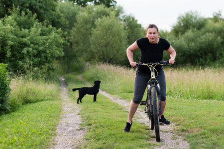 A woman on a bicycle pauses on a country path, her black dog waits patiently nearby. The scene evokes a sense of peace and outdoor activity.の写真素材