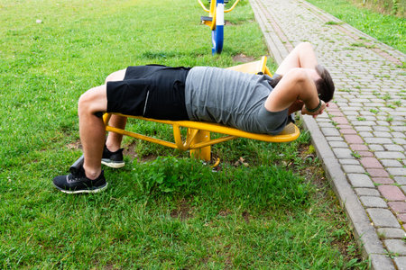 A man performs sit-ups on yellow outdoor gym equipment. He's wearing athletic clothing and is exercising in a grassy park setting near a paved path.の写真素材