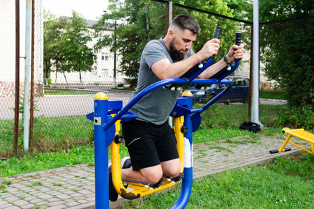 A bearded man performs core exercises on a public outdoor gym machine. He's kneeling, focused on his workout in a green park environment.の写真素材