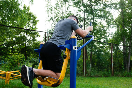 A person engages in a back workout using an outdoor fitness machine. The setting is a park with green grass and trees. The equipment is simple and sturdy.の写真素材