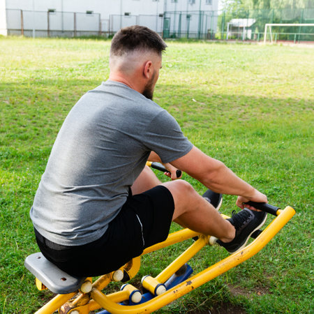 A man uses an outdoor fitness machine, focusing on a rowing motion. The scene is set in a grassy park-like area.の写真素材