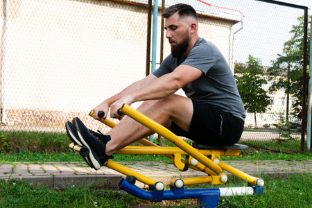 A bearded man uses outdoor gym equipment, focusing on a chest press. He's dressed in athletic wear and appears determined and healthy. The setting is a sunny park.の写真素材