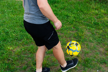 A person wearing athletic shorts and shoes skillfully juggles a yellow and black soccer ball on a lush green lawn. The scene suggests leisure and physical activity.の写真素材