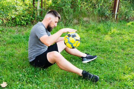 A bearded man sits on a grassy field, carefully examining a yellow and black soccer ball. He's dressed in athletic attire, suggesting a break from sports activity.の写真素材