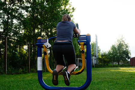 A woman uses an outdoor exercise machine in a park setting. The machine is blue and yellow, and she is wearing athletic clothing. The grass is green and well-maintained.の写真素材