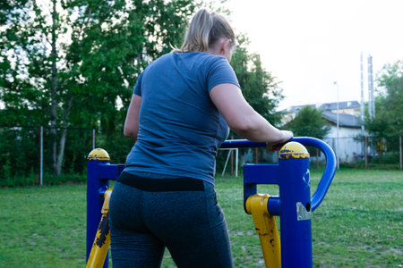 A woman uses a public outdoor fitness machine. She is wearing a gray shirt and dark gray leggings....