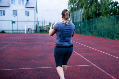 A woman in sportswear walks on a red sports court. She carries a ball and is seen from behind, heading toward a goal or net. The court is outdoors.の写真素材