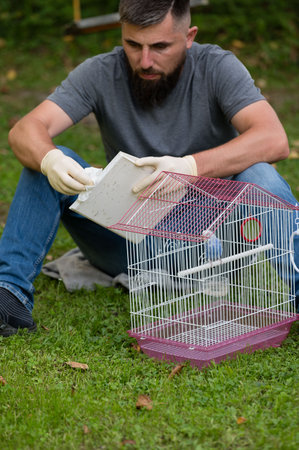 A bearded man sits on the grass, wearing gloves, cleaning a small birdcage. The cage is pink and white. The scene is peaceful and outdoors.の写真素材